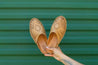 A hand holding a pair of natural leather slip-on sandals with hand-woven diamond centerpiece in front of green background