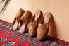 Two pairs of natural leather slip-on sandals with hand-woven diamond centerpiece on red rug propped against wall